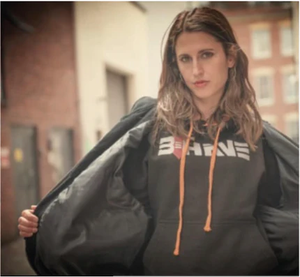 Female model wearing a black behave hoodie with a brand logo, standing outdoors.